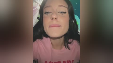 Media: Video of a young woman with fair skin, long black hair, wearing a pink T-shirt with a partially visible text. She has a septum piercing and is making a silly face. Background shows a green and white room.