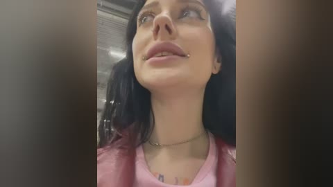 Media: Video of a young woman with long black hair, fair skin, and a small nose piercing. She wears a pink top, looking upward, possibly in a car. Background is blurry.