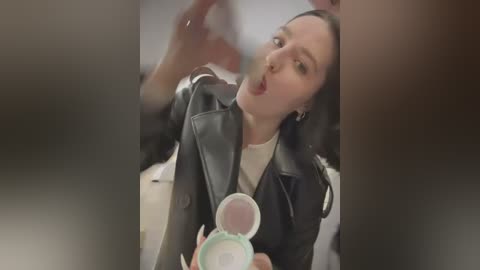 Media: Video of a woman in a black leather jacket, with a surprised expression, holding a green container, indoors.