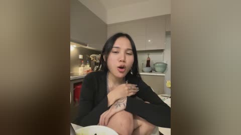 Media: A video of an Asian woman with long black hair, wearing a black top, sitting on a kitchen counter, holding a bowl, with a tattoo on her arm, in a modern kitchen with beige cabinets and white walls.