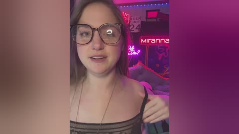 Media: A video of a young woman with glasses, light skin, and shoulder-length brown hair, wearing a black top and necklace, smiling, in a dimly lit room with neon signs, including \"MIRANNA\" and \"BRAVE.\