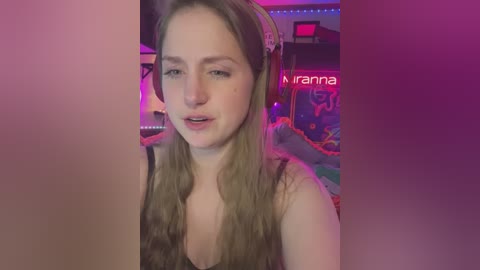 Media: Video of a young woman with long blonde hair, wearing headphones, sitting in a dimly lit room with neon lighting. She has a light complexion and is wearing a black top.