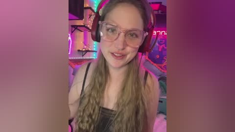 Media: Video of a young woman with long, wavy blonde hair, wearing clear glasses and a black tank top, sitting in a colorful, cluttered room with neon lights and a gaming setup.