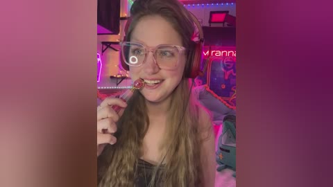 Media: A video of a young, fair-skinned woman with long, light brown hair and large, clear glasses, wearing headphones, licking a lollipop. She's smiling and has a casual, relaxed demeanor. The background is a dimly lit, colorful room with neon lights.