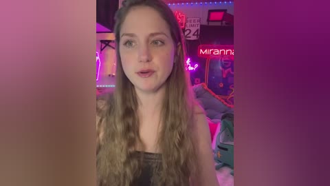 Media: A video of a young woman with long, wavy blonde hair, fair skin, and a slender build, wearing a black top. She is indoors with neon lights and a colorful, cluttered background.