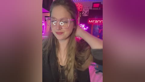 Media: Video of a smiling, fair-skinned woman with long blonde hair, wearing clear glasses and a black top, sitting in a dimly-lit, neon-lit room with various signs and a \"Mirrann\" watermark.