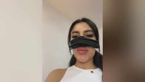 Media: Video of a woman with long black hair, wearing a black face mask and white top, against a plain white background.