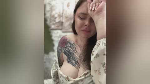 Media: Video of a young woman with fair skin, long brown hair, and large breasts, wearing a floral off-shoulder dress. She has a tattoo of a black and gray rose on her right shoulder. She is indoors, looking down, her face showing a serene expression.