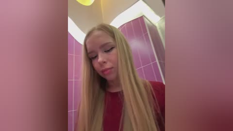 Video of a blonde, fair-skinned woman in a red top, standing in a pink-tiled bathroom, looking downward with a contemplative expression.