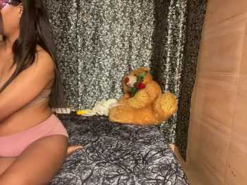 Media: Video of a curvy woman with medium-dark skin, wearing pink panties and a black bra, sitting on a bed with a beige teddy bear and black floral-patterned bedspread.