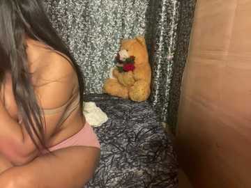 Media: Video of a woman with long black hair, wearing pink lingerie, sitting on a bed with a teddy bear and a red rose. Background features a patterned curtain and a wooden wall.