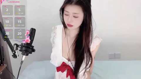 Media: Video of a slender, fair-skinned Asian woman with long black hair, wearing a white dress with a red bow, singing into a microphone in a bright, minimalistic room with a calendar on the wall.