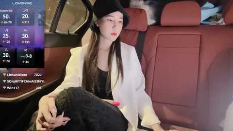 Media: Video of a woman with long, straight hair wearing a black hat, white blazer, and black pants, sitting in a red leather car seat. Background shows a dark, possibly night-time setting.