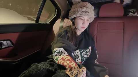 Media: Video of a young woman wearing a black sweater with a colorful geometric pattern, a knit beanie, and a blanket, sitting in the back seat of a red car with a snow-covered exterior.