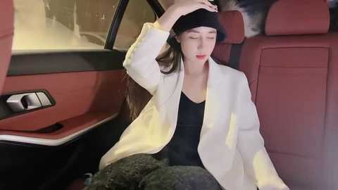 Media: Video of a young woman with long brown hair, wearing a white blazer and black beanie, sitting in a modern car with red and black interior.