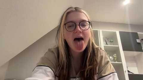 Media: Video of a young Caucasian woman with long blonde hair, wearing glasses and a beige hoodie, sticking out her tongue in a playful pose, indoors with white cabinets and a lens flare in the background.