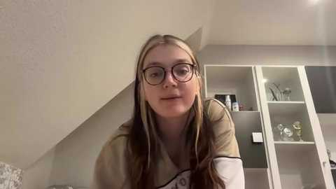 Media: Video of a young woman with long, straight blonde hair, wearing glasses, and a beige shirt, standing in a modern, minimalist room with white cabinets and shelves.