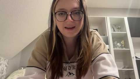 Media: Video of a fair-skinned woman with long brown hair and glasses, wearing a beige sweatshirt with a mountain graphic, smiling, leaning forward, in a modern, white-walled bedroom with built-in shelves and decorative items.