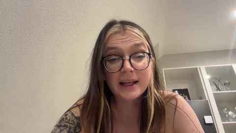 Media: Video of a young woman with fair skin, glasses, and brown hair, wearing a sleeveless top, leaning against a beige wall. Background features a white-framed mirror and a few decorative items.