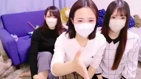 Media: Video of three Asian women in masks, sitting on a purple couch, wearing white tops and plaid skirts, in a room with blue curtains.