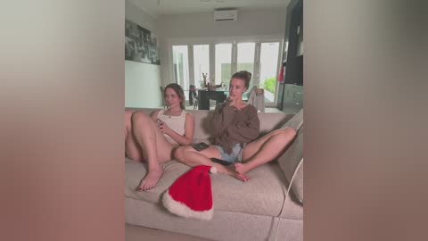 Media: Video of two young women in a modern living room, one wearing a red Santa hat, the other in a brown sweater, sitting on a beige couch, chatting.