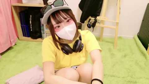 Media: A video of a young Asian woman with light skin, wearing a yellow shirt, headphones, and a pink face mask, sitting on a green carpet in a cluttered room with bookshelves and hanging clothes.