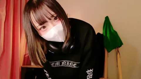 Media: Video of a young woman with straight brown hair, wearing a black sweatshirt with white text, a white face mask, and a green towel hanging behind her.