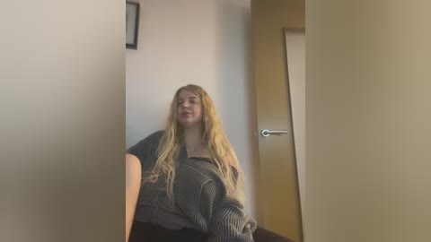 Media: Video of a blonde woman with long hair, wearing a gray sweater, sitting in a hallway with beige walls and a wooden door.