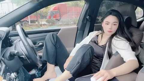 Media: Video of a young Asian woman with long black hair, wearing a black top, white cardigan, and black pants, sitting in a car with a white seat cover, outside a red truck, blurred background.