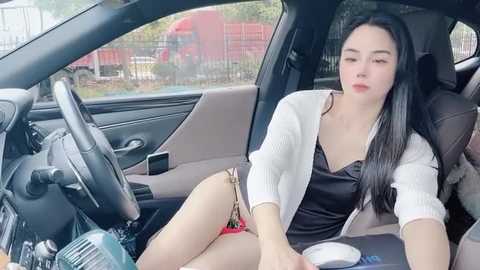 Media: A video of a young East Asian woman with long black hair, fair skin, and wearing a black camisole and white cardigan, sitting in a car, looking relaxed.