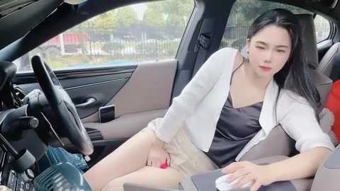 Media: A video of a young East Asian woman with long black hair, wearing a black top and white blazer, sitting in a car, holding a red lipstick.