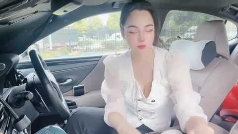 Media: Video of a woman with fair skin and dark hair, wearing a white blouse, sitting in a car with beige seats and a red object beside her.