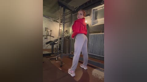 Media: Video of a young woman with light skin and brown hair in a red jacket and gray leggings, working out in a dimly lit gym with a weight machine and a radiator.