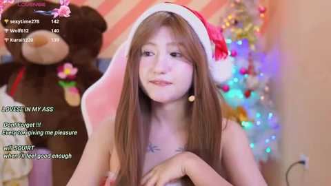 Media: Video of a young Asian woman with long brown hair, wearing a red Santa hat and nothing else, standing in front of a Christmas tree, with text overlay.