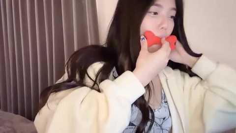 Media: A video of a young Asian woman with long black hair, wearing a light cream cardigan, playing with a red toy. She is indoors, against a beige wall with vertical blinds.