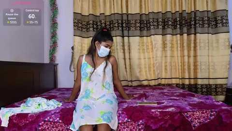 Media: Video of a young woman with dark skin and long hair in a floral dress, sitting on a bed with a pink and purple quilt. She wears a white face mask and is in a bedroom with beige curtains.