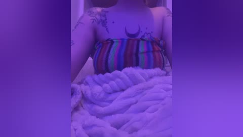 Media: Video of a fair-skinned woman with a crescent moon tattoo on her chest, wearing a colorful striped tube top, covered with a fluffy white blanket, set against a purple backdrop.