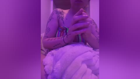 Media: A video of a tattooed person, wearing a colorful striped shirt, holding a fluffy white blanket under a purple light.