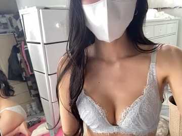 Media: A video of a woman with long black hair, wearing a white face mask, light gray lace bra, and beige shorts, standing in a cluttered bedroom with white furniture and a mirror.