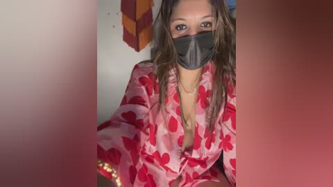 Media: Video of a woman with medium-brown skin, wearing a red floral-patterned robe, black face mask, and wet hair, sitting indoors with a blurred background.