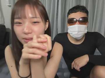 Media: Video of an Asian woman with straight brown hair and light skin, wearing a black tank top, sitting on a couch. Next to her is a man in a black long-sleeve shirt, white face mask, and dark sunglasses, also wearing a mask.