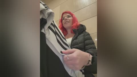 Media: A video of a smiling woman with vibrant red hair, wearing a black puffer jacket and a black shirt, holding a striped garment in a beige-tiled bathroom.