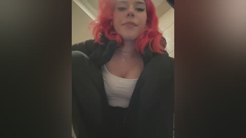 Media: A video shows a young woman with bright red, curly hair, wearing a black jacket over a white top, seated in a public restroom with beige tiles in the background.