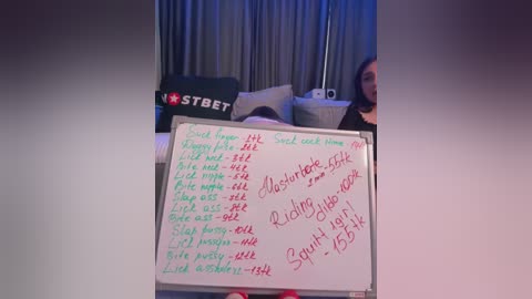 Media: Video of a woman in a dark room, holding a whiteboard listing various sexual acts, including \"Rimming,\" \"Fisting,\" and \"Anal.\" Background includes a couch and a \"STET\" sign.