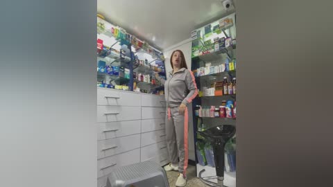 Media: Video of a woman in a gray lab coat standing in a well-lit pharmacy with neatly organized shelves and drawers, holding a clipboard.