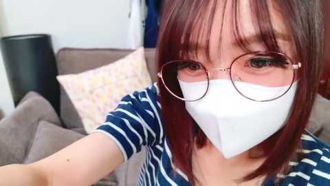 Video of an Asian woman with light skin, straight brown hair, and round glasses, wearing a white surgical mask, a blue and white striped shirt, and a beige couch with patterned pillows in the background.