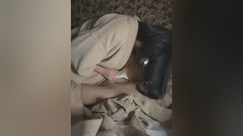 Media: Video of a person with black hair and a beige blanket covering their head and body, lying on a bed with a textured brown blanket. The room is dimly lit.