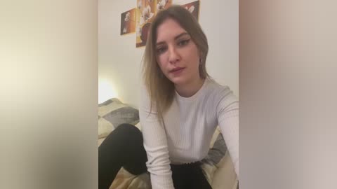 Media: Video of a young Caucasian woman with light skin, shoulder-length blonde hair, and a white ribbed long-sleeve top. She sits on a beige couch, wearing black leggings. The background shows a white wall with framed pictures and a warm light source.