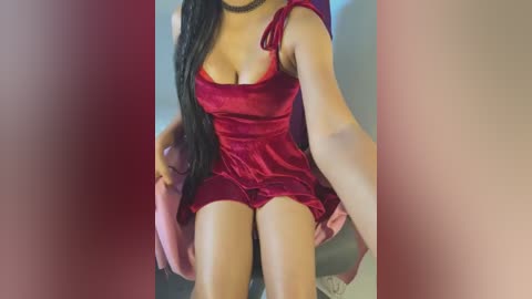 Media: Video of a woman in a red velvet mini-dress, sitting on a pink couch, showing off her medium-sized breasts and toned legs. The background is blurred, with a soft pink and beige gradient.