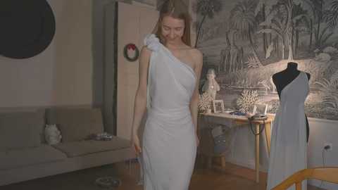 Media: Video of a slender, fair-skinned woman with long blonde hair in a white, one-shoulder dress, standing in a modern, minimalist room with a grey couch, a large black and white mural, and a mannequin dressed in a white dress.